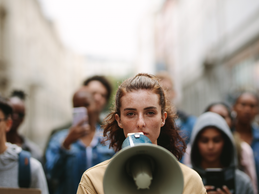 Eine Frau hat ein Megafon vor dem Gesicht. Hinter ihr sind Menschen zu sehen, die in ihre Richtung mitlaufen